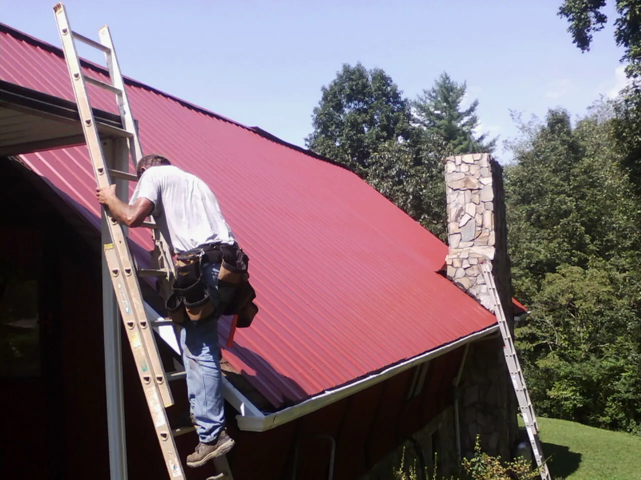 Roofer on a ladder beside a red metal roof and stone chimney — Soffit & Fascia Repair by Roofing Brothers in Caroleen